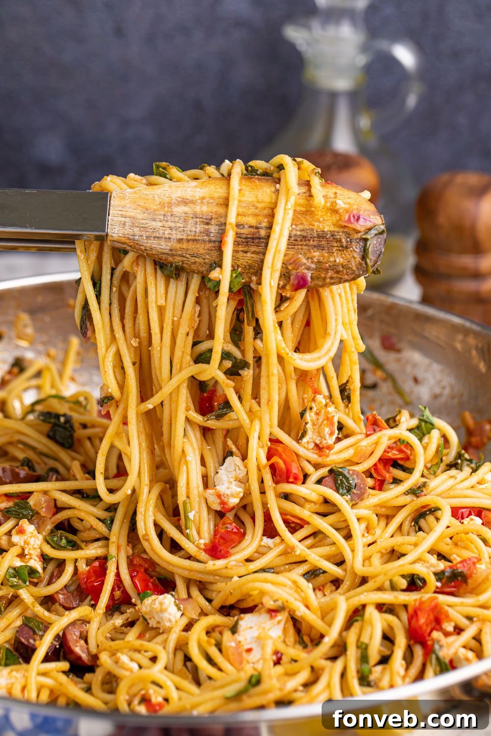Greek Spaghetti using tongs to lift some of the pasta and veggies out of the pan, demonstrating its readiness to serve.