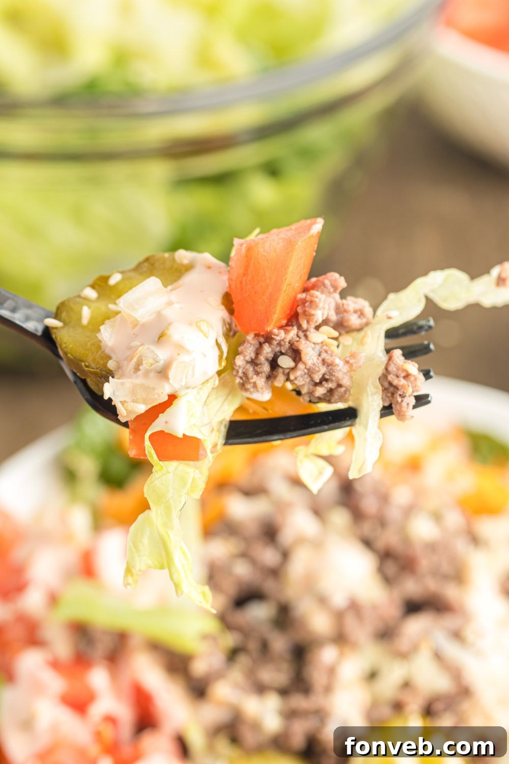 Big Mac Makeover Salad 4 Close-up view of Big Mac Salad in a bowl on a table, with a fork lifting a portion of the salad to show the layers of ingredients.