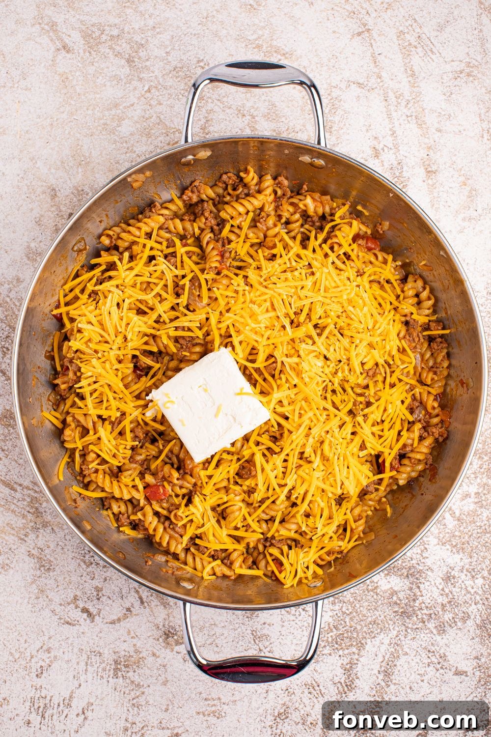 A skillet of Taco Macaroni and Cheese with cubes of cream cheese and a mound of shredded cheddar cheese melting on top, creating a rich sauce.