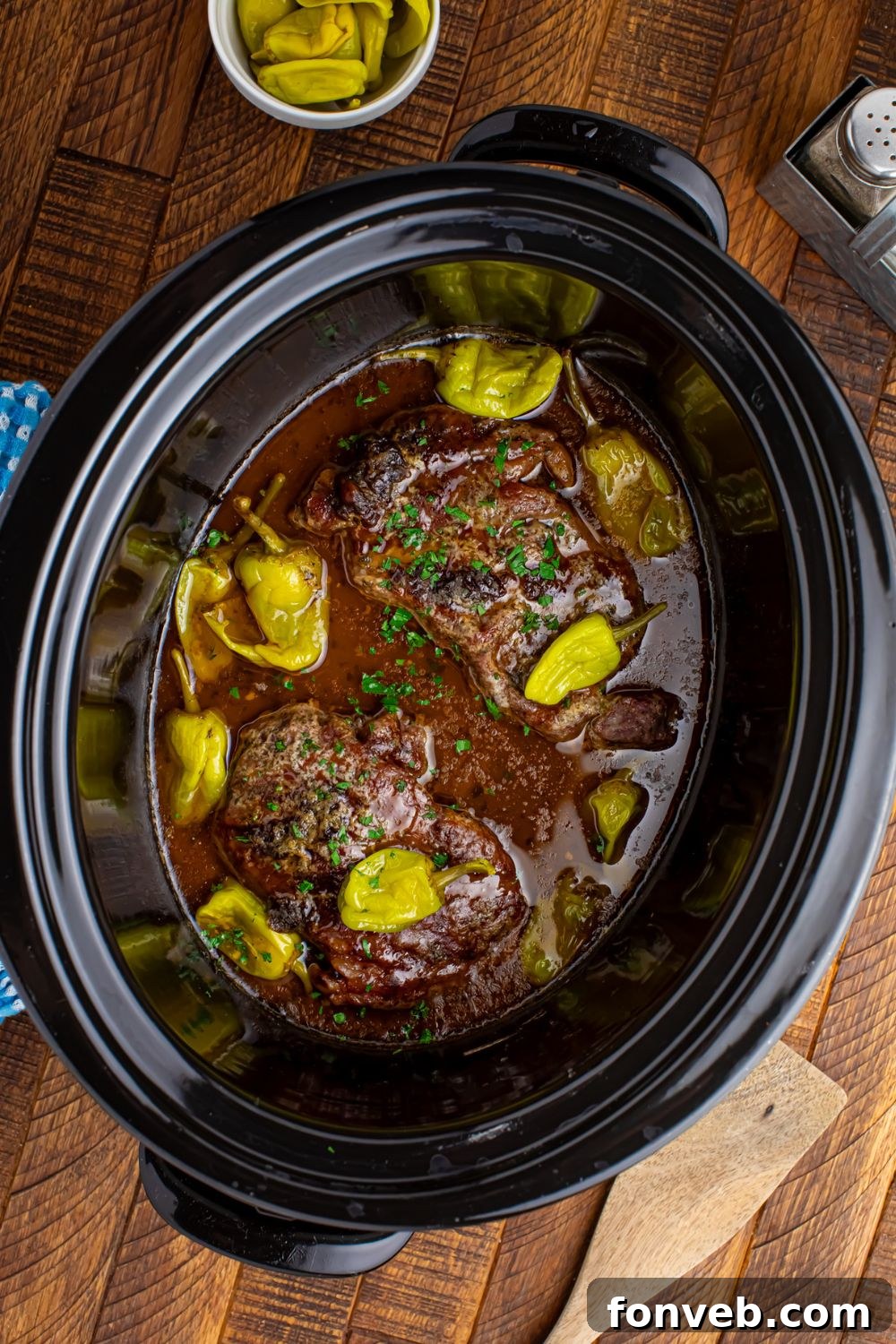 Slow Cooker Mississippi Ribeye Steaks in crock po on wooden table wih peppers in a bowl tto the side