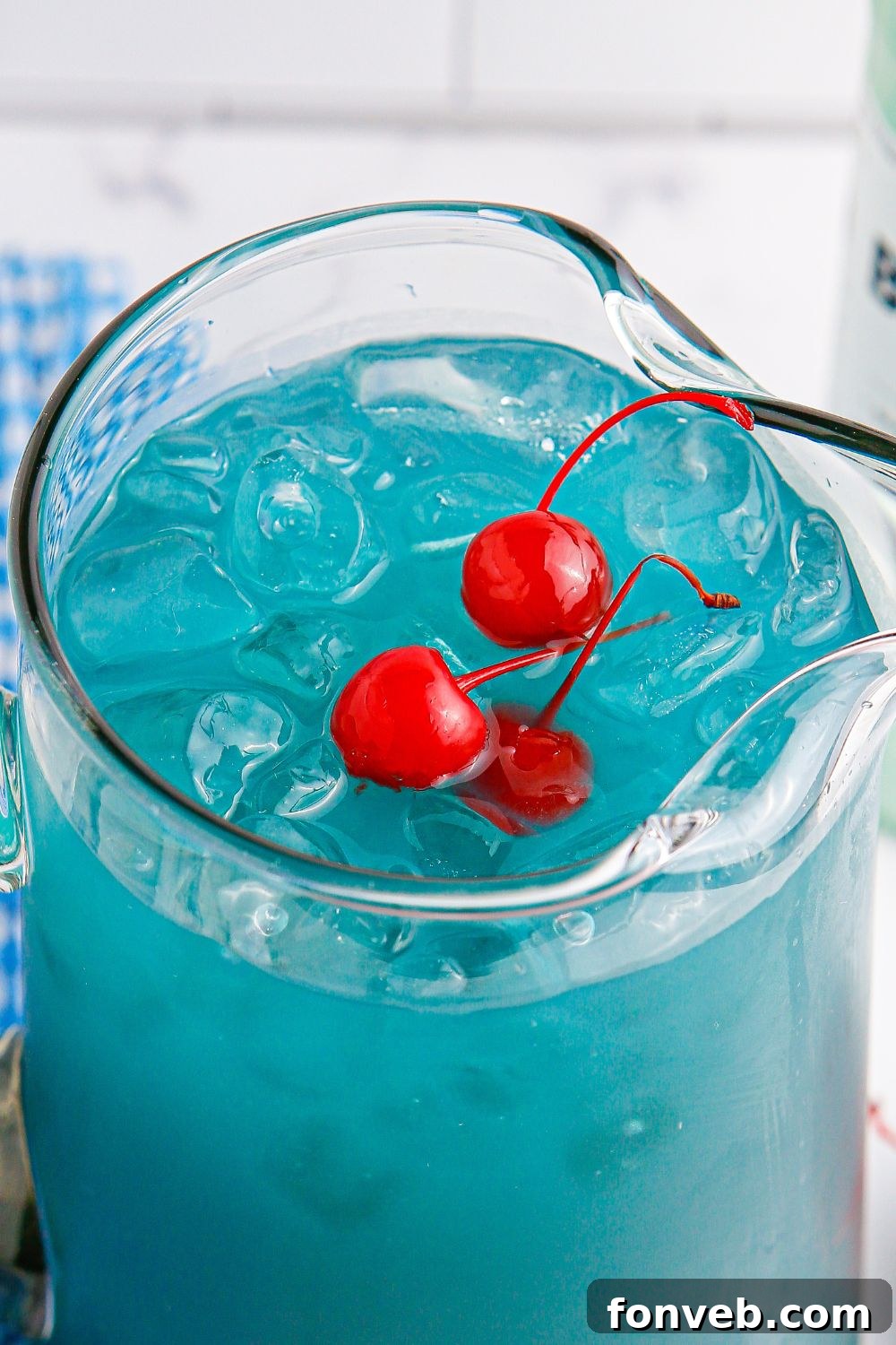 A glass pitcher filled with shimmering Blue Hawaiian Punch and ice, adorned with vibrant red maraschino cherries on top.