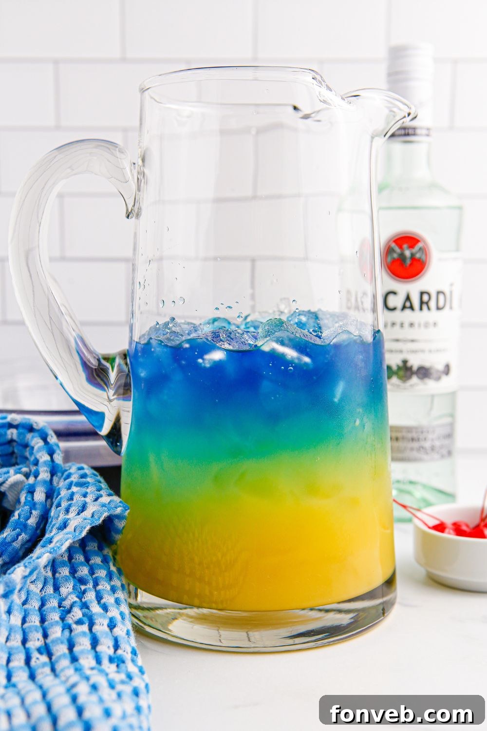 Preparing the Blue Hawaiian Punch by carefully layering the vibrant ingredients into a large glass pitcher on a table.