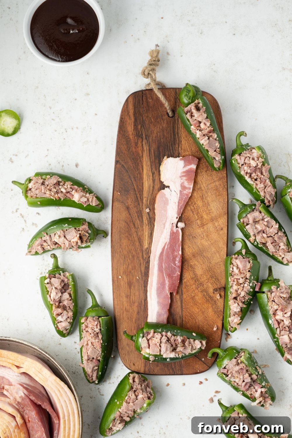 slice of raw bacon on cutting board with stuffed jalapenos sitting around it on table