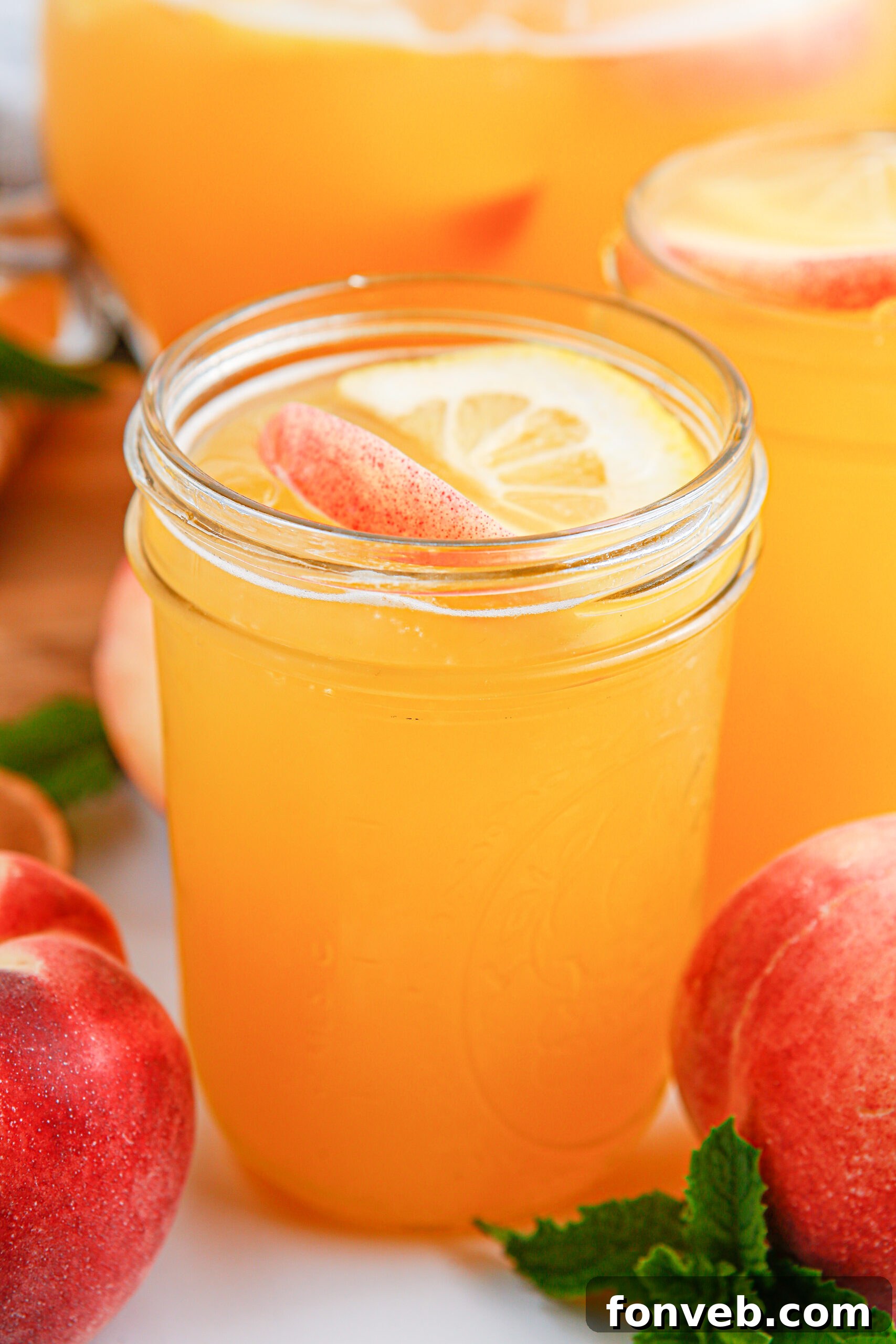 Sun-Kissed Peach Lemonade 2 Peach Lemonade in a mason jar glass garnished with peach slice and orange slice. Fresh peaches around the glass