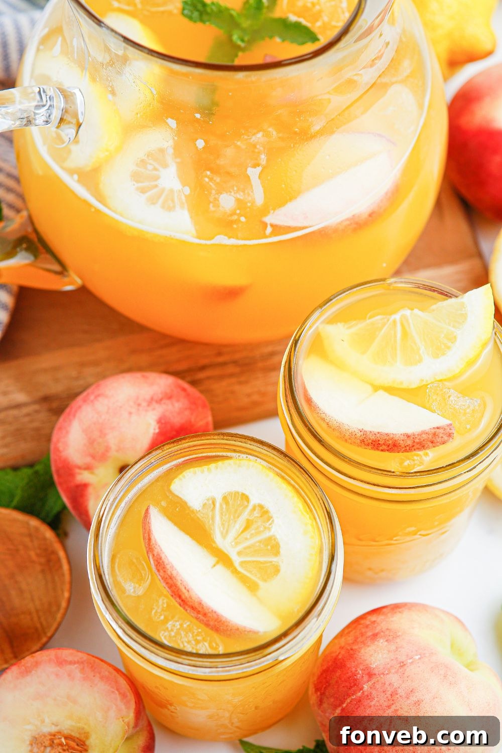 Sun-Kissed Peach Lemonade 4 large pitcher with peach lemonade on table. Mason jars full of lemonade and fresh peaches scattered on table