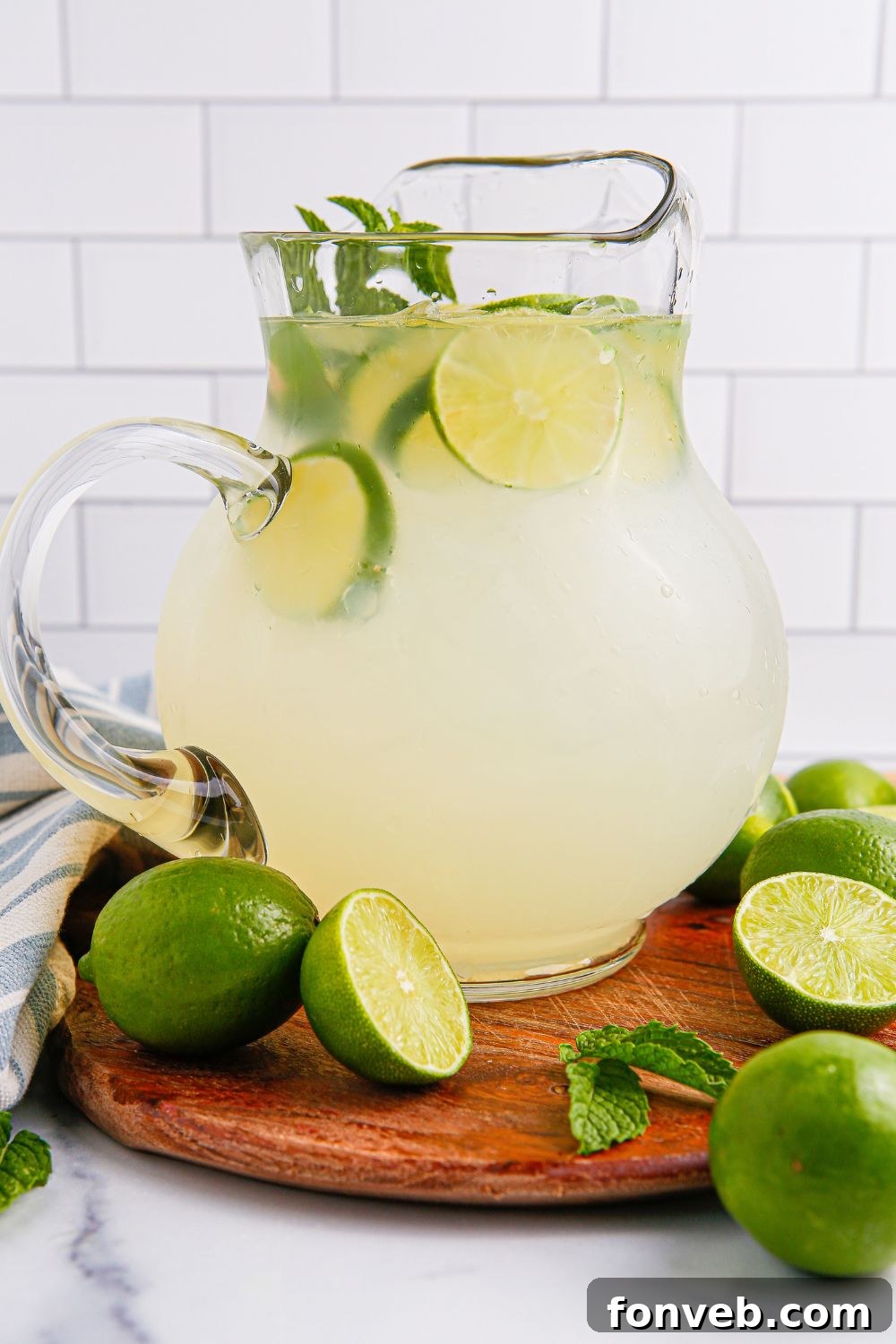 side view of limeade in a glass pitcher sitting on a wooden cutting board and limes placed around the pitcher