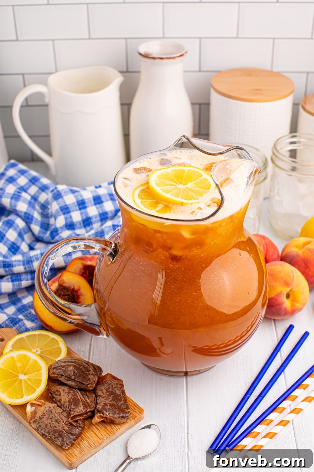 large pitcher of peach sweet tea on table with straws, sugar, lemon slices and more around the pitcher