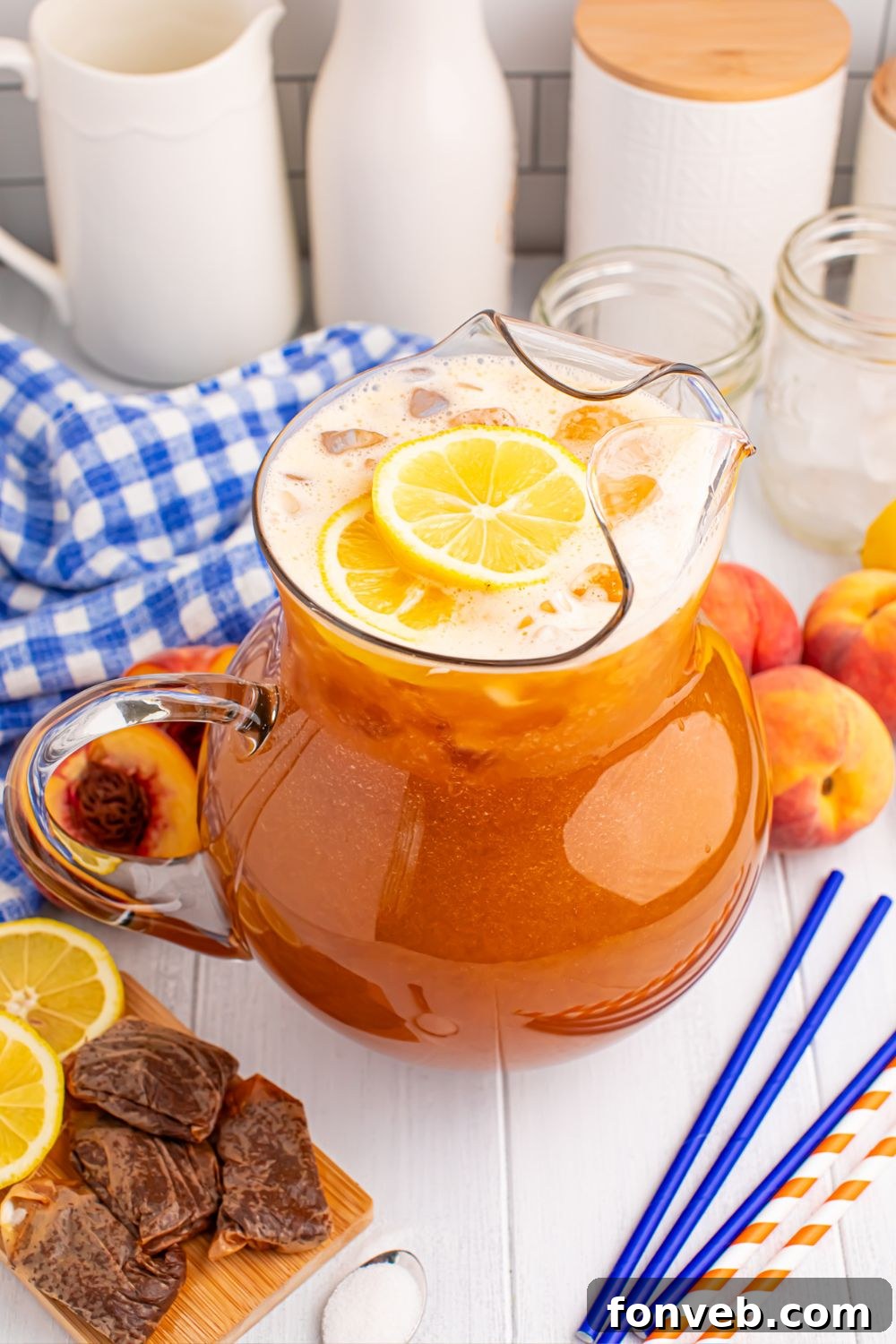 peach tea on the table with wet tea bags, slices of fruit, straws, sugar, and a blue checked towel around the container