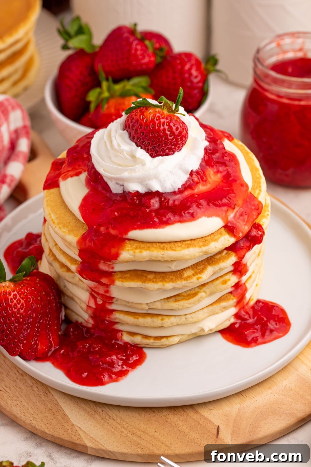 A stack of Strawberry Cheesecake Pancakes, topped with whipped cream and a whole strawberry, with a bowl of fresh berries in the background.