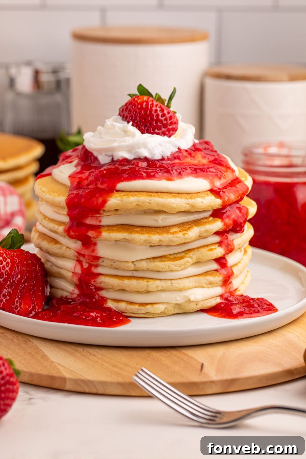 A plate of Strawberry Cheesecake Pancakes on a table with baking ingredients blurred in the background, a fork, and fresh strawberries.