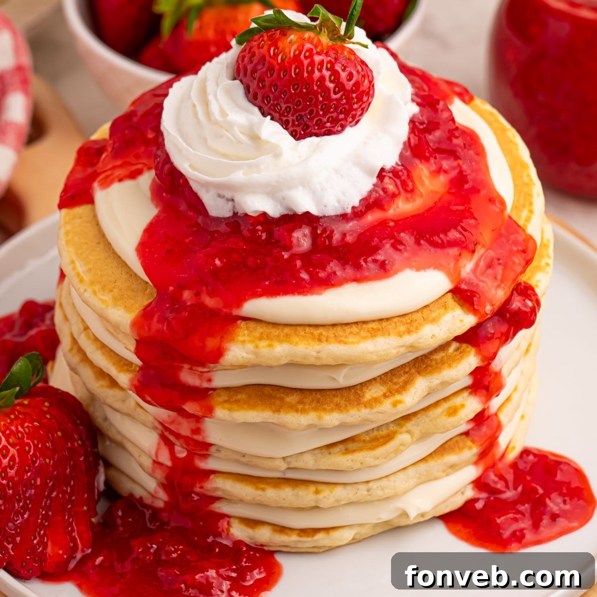 Tall stack of Strawberry Cheesecake Pancakes topped with whipped cream and a single fresh strawberry, on a white plate.