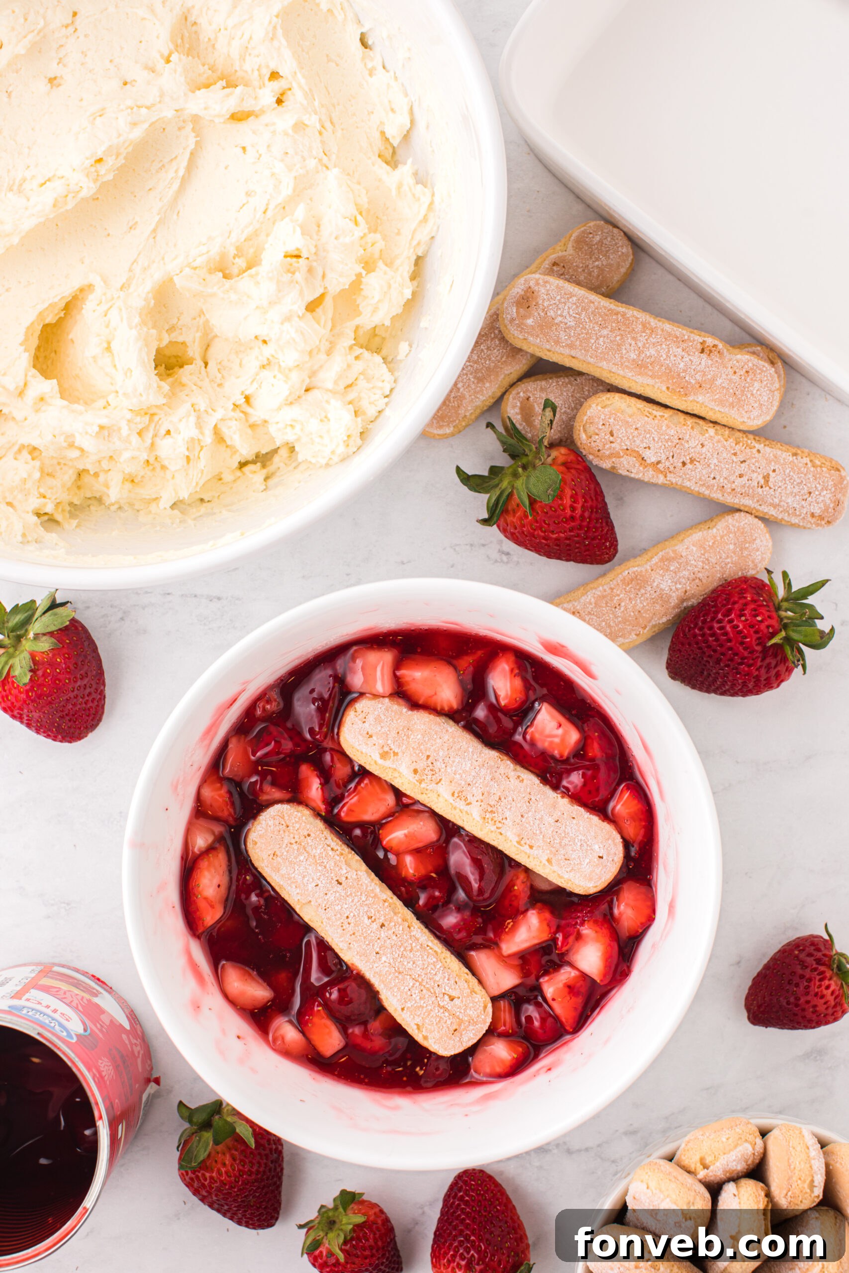 ladyfingers in a strawberry mixture and a bowl of mascarpone filling behind it 