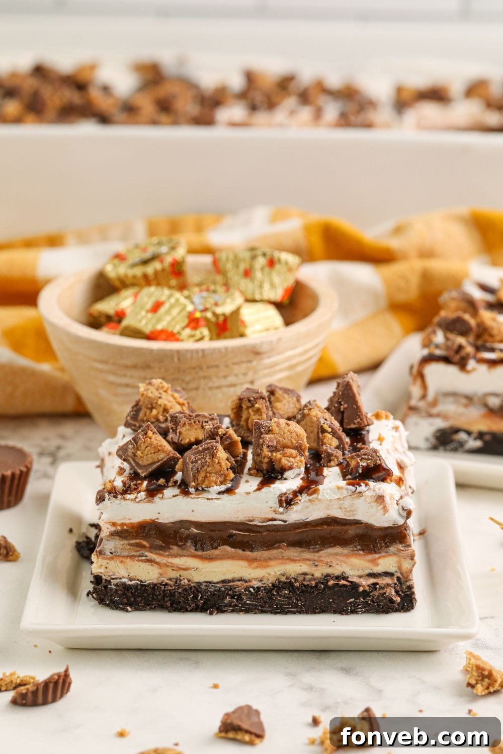 Peanut Butter Chocolate Sweet Stack 15 Reeses Lasagna sliced on table with a bowl of candies in background and pan behind it on table