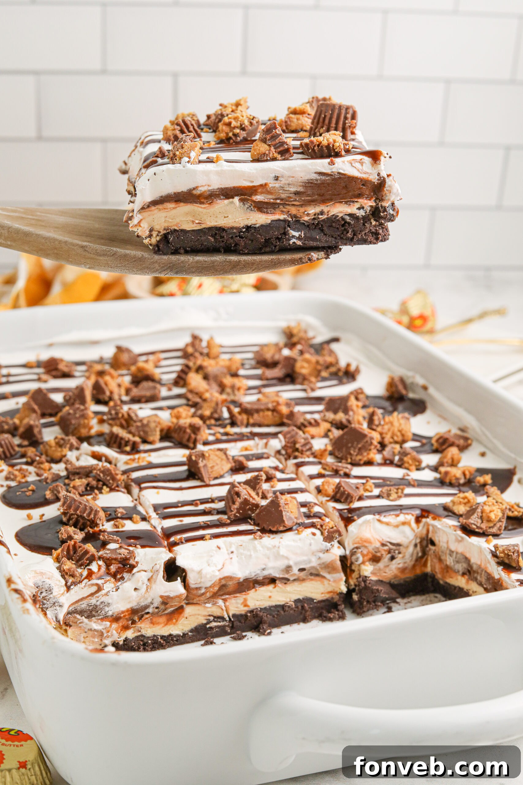 Peanut Butter Chocolate Sweet Stack 17 Reeses lasagna in a white baking pan with a piece being lifted up