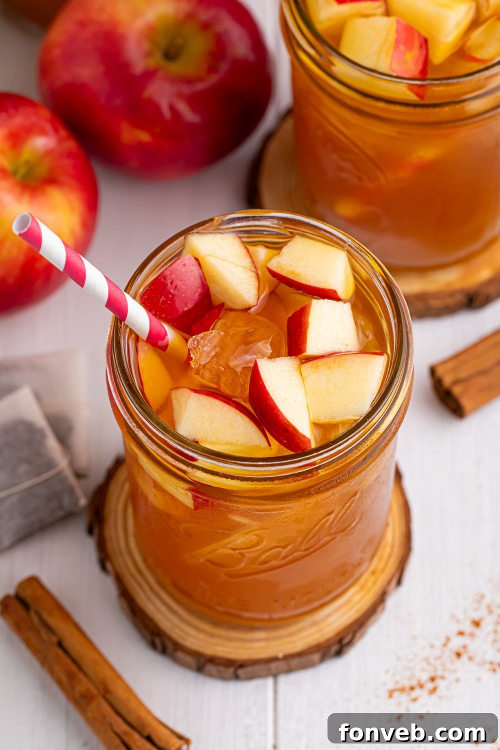 mason jar with an apple tea sitting on it on a wooden coaster and other glasses and apples spread over the table with a sprinkle of cinnamon on table 