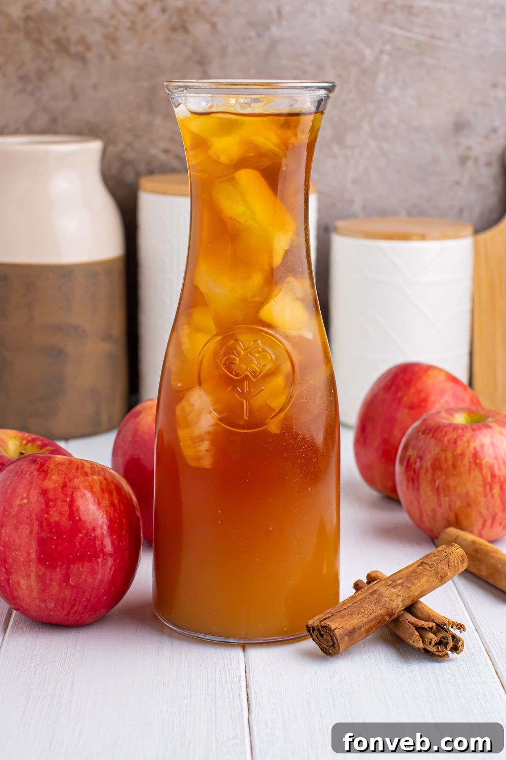 cider tea in a pitcher with cinnamon sticks and whole apples around it showing this is an apple cider iced tea