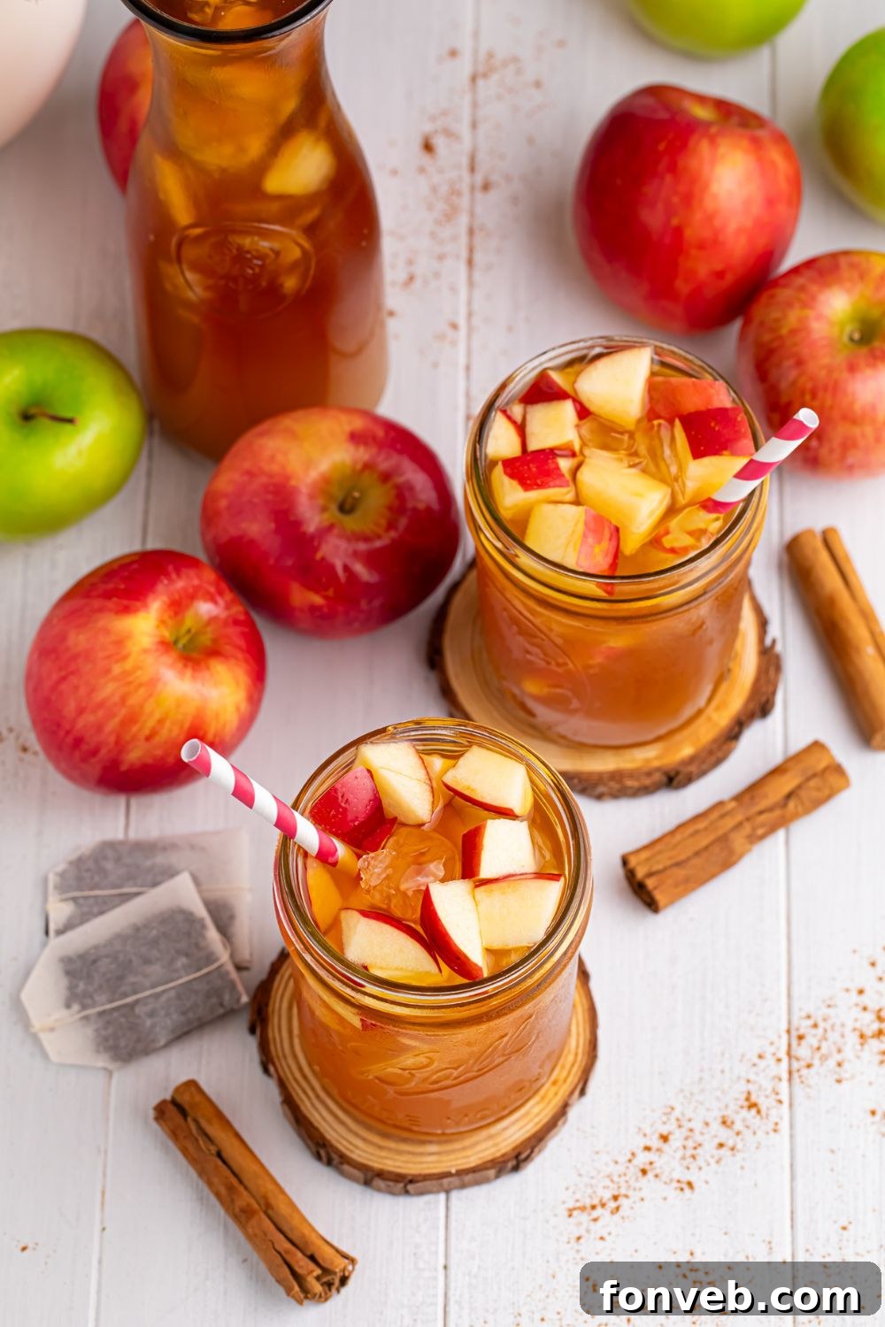 Apple Cider Iced Teas on table in two glasses, a large pitcher, with tea bags, cinnamon sticks and apples all over table