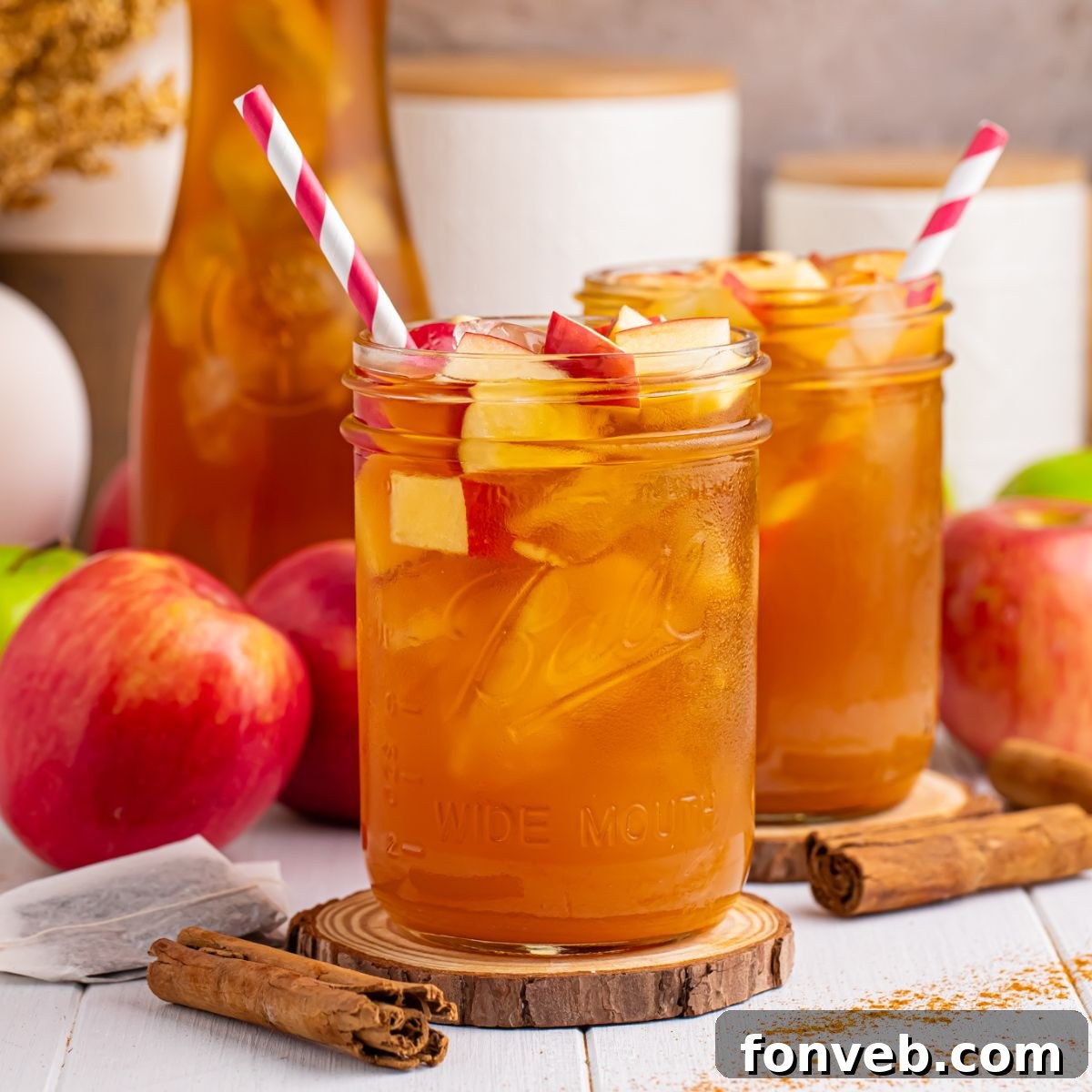 Apple Cider Iced Tea glasses on table with apples and cinnamon sticks around glasses and a red and white striped straw in each mason glass