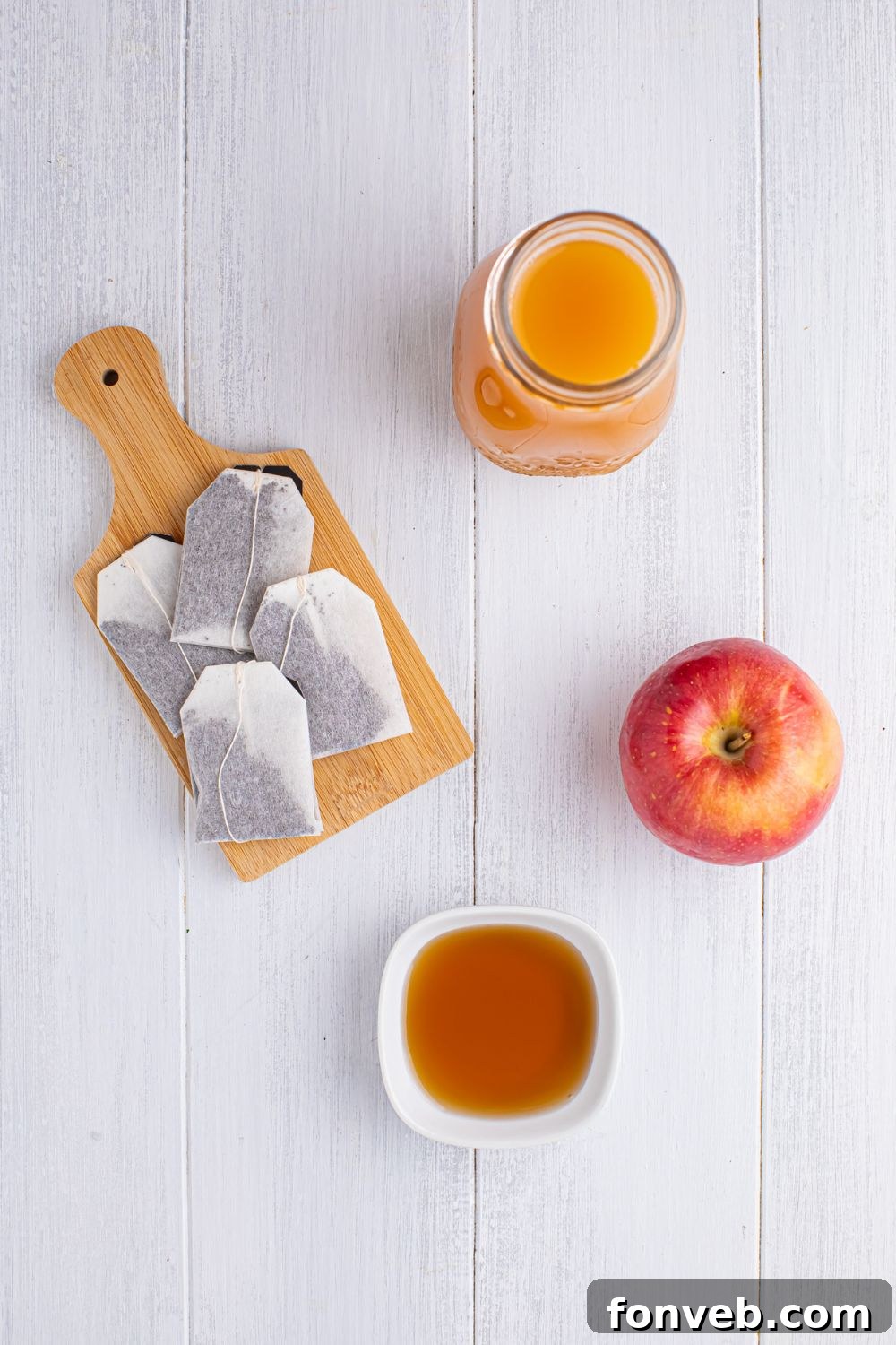 ingredients for making apple cider iced tea on a white table in single serve bowls: tea bags on a cutting board, mug of apple cider, whole apple 