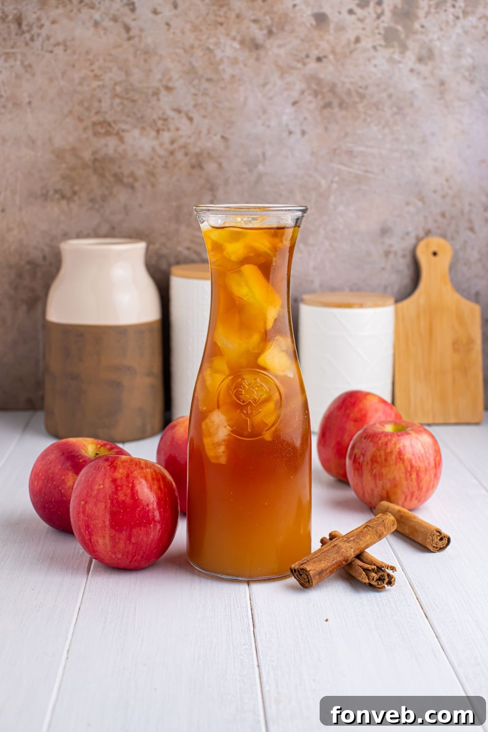 small pitcher of apple cider iced tea on table with apples and cinnamon sticks around it, and jars and cutting board in the background 
