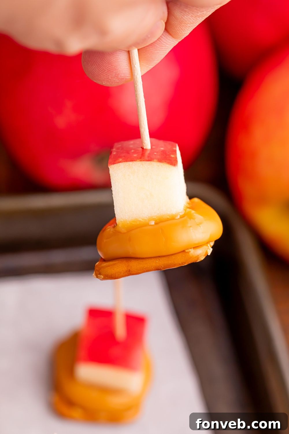 Caramel Apple Pretzel Bite being held in hand with toothpick with tray of more on table and apples around the tray 