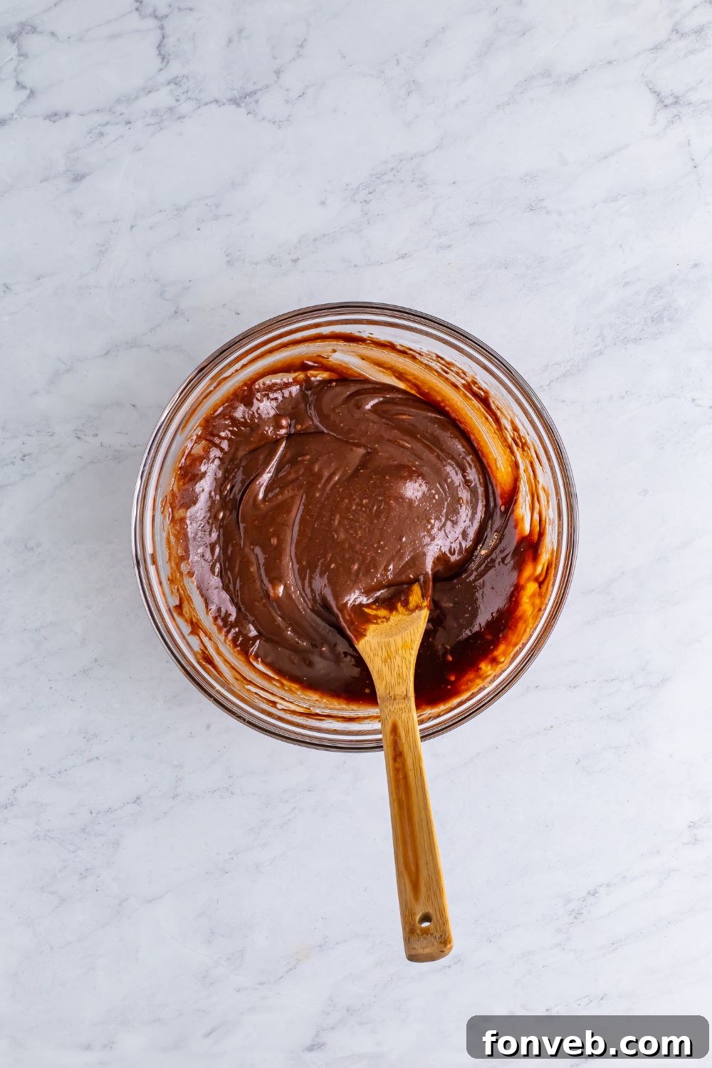 A glass bowl filled with wet brownie batter, a wooden spoon resting inside, showcasing its rich, chocolatey texture