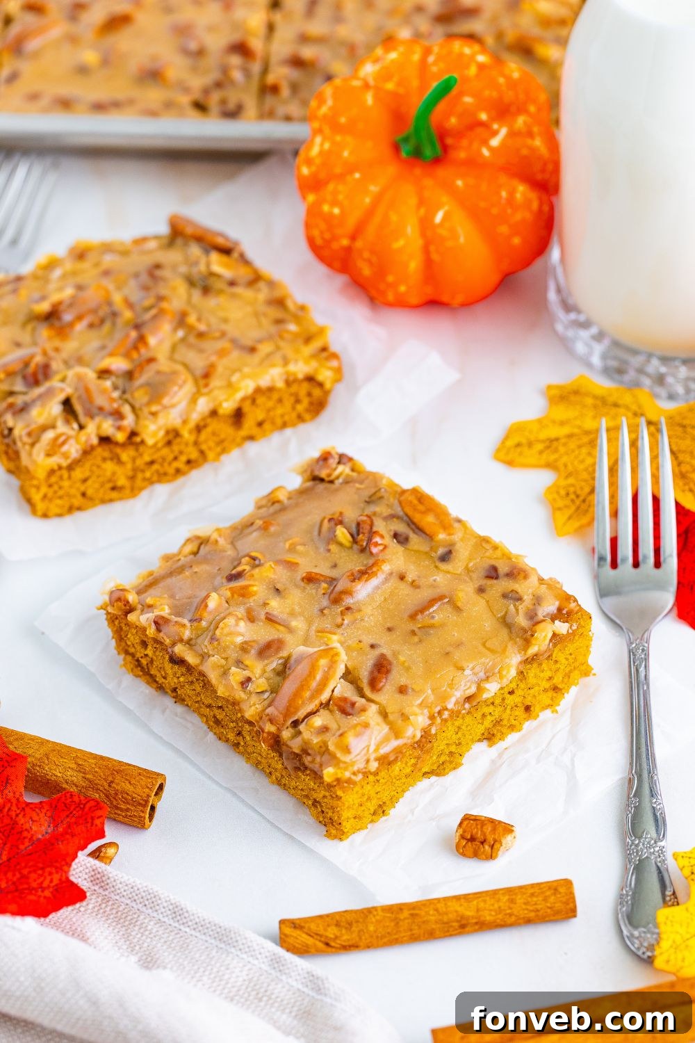 Perfectly cut squares of Pumpkin Snack Cake with Praline Icing arranged on a table, accompanied by a fork, a decorative faux pumpkin, and a refreshing glass of milk, evoking a cozy fall scene.