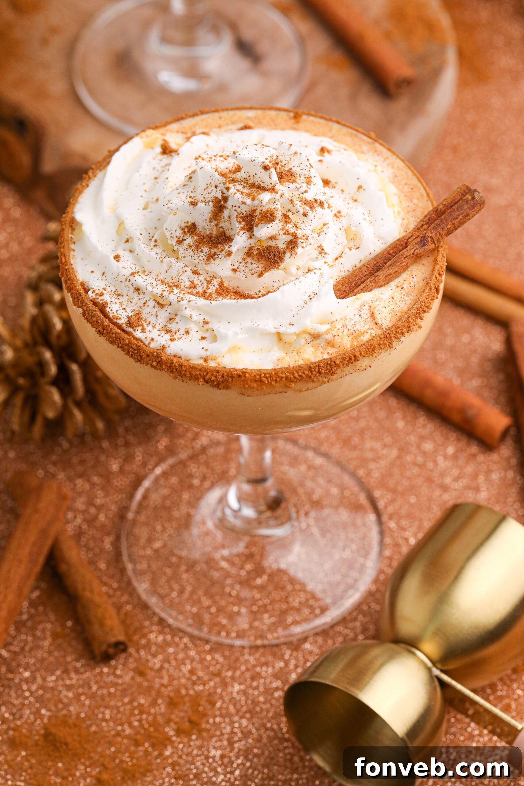 A festive Pumpkin Pie Martini in a clear glass, garnished with a cinnamon stick, sits on a beautifully decorated holiday table, evoking warmth and autumn cheer.