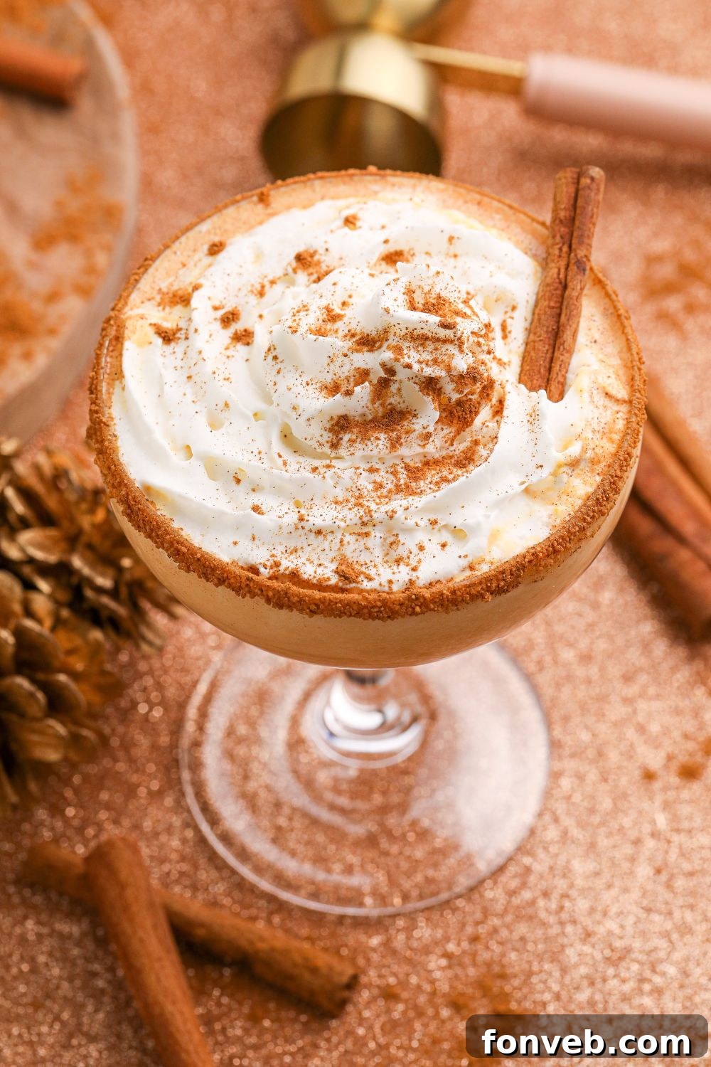 A close-up of a Pumpkin Pie Martini, beautifully topped with whipped cream and a cinnamon stick, sitting on a table with more cinnamon sticks scattered around.