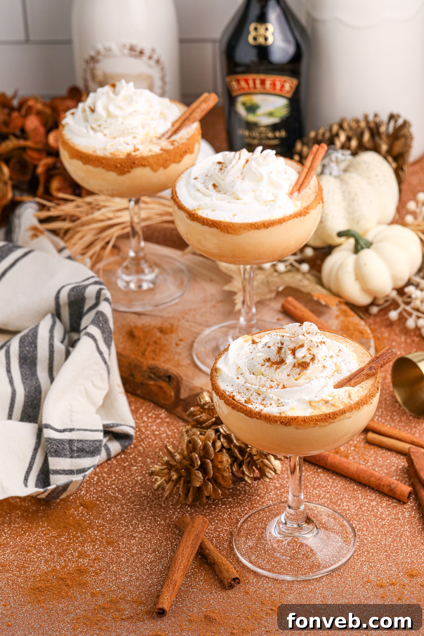 Two glasses of pumpkin martinis are placed on a table with bottles of alcohol in the background, surrounded by fall decorations, setting a cozy, autumnal scene.