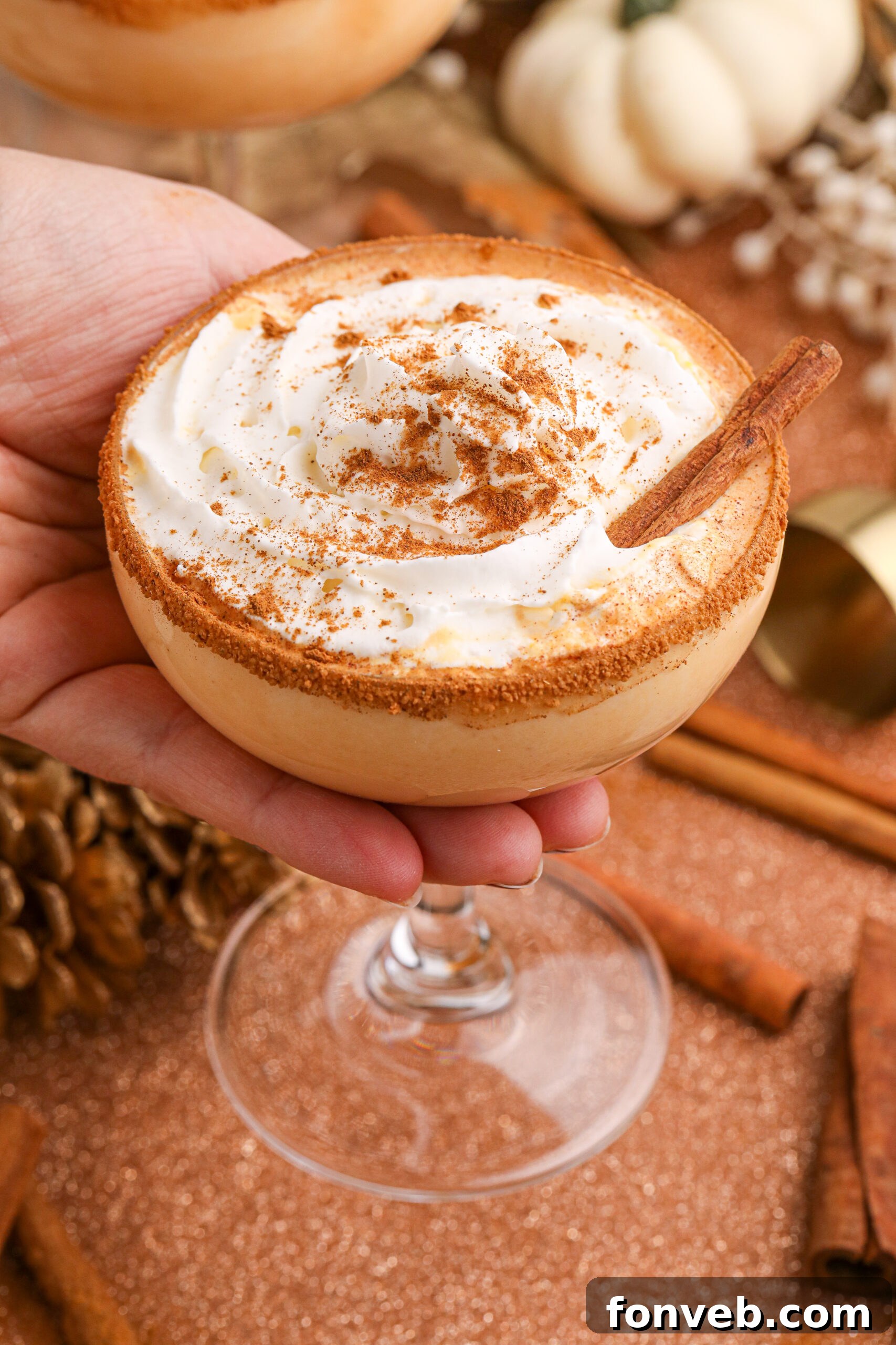 A hand gently holds up a glass of Pumpkin Pie Martini, showcasing its creamy texture and inviting amber hue against a softly blurred background.