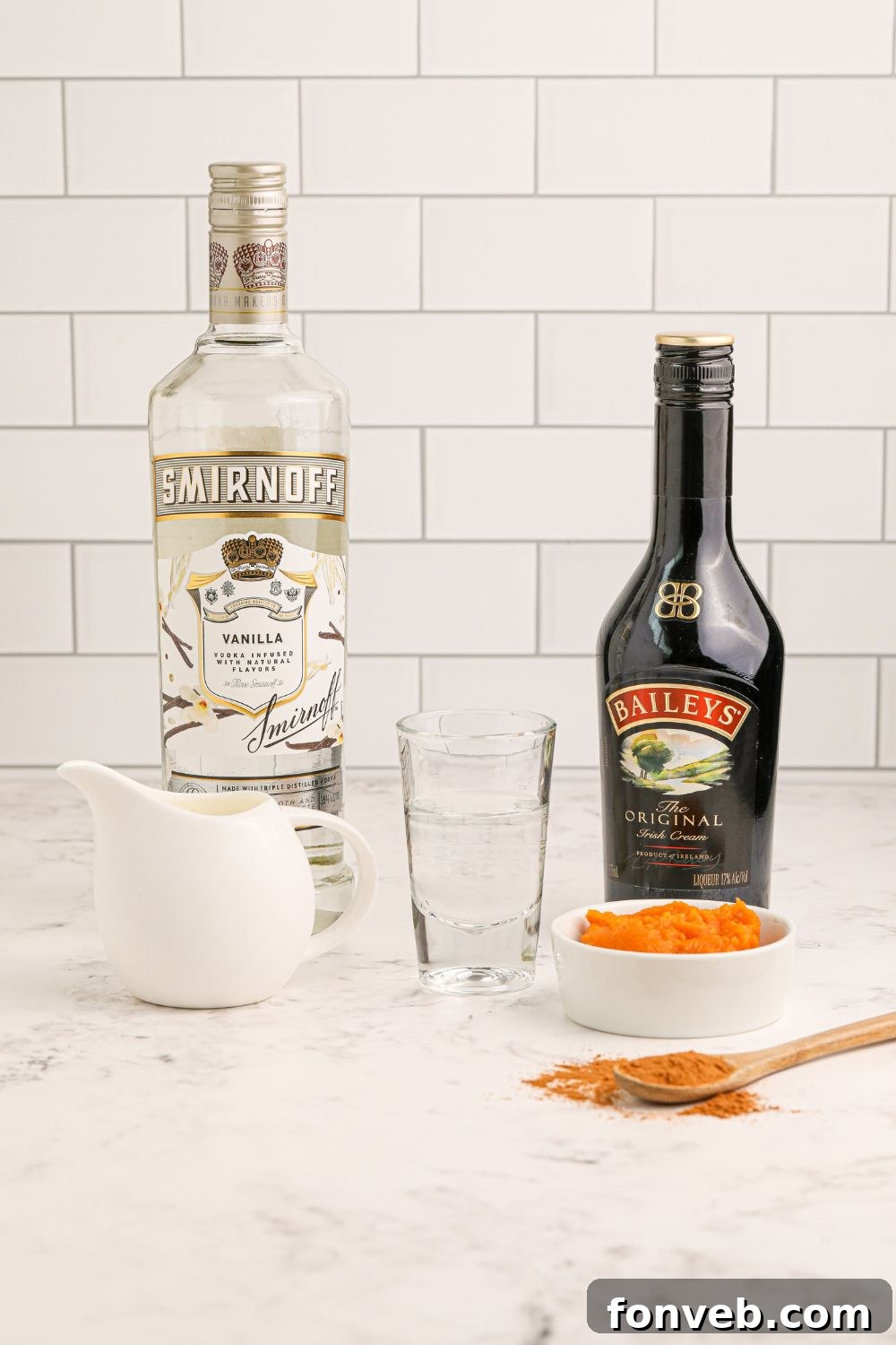 Individual ingredients for a Pumpkin Pie Martini laid out: Baileys Irish Cream, vanilla vodka, heavy cream, pumpkin puree, and pumpkin pie spice, ready for mixing.