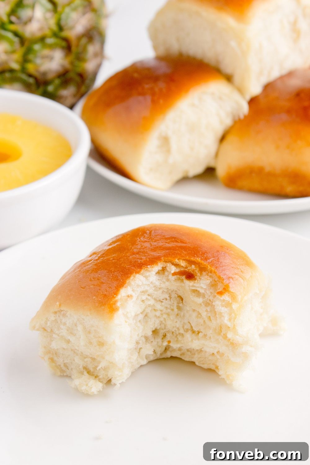 Tropical Dinner Buns 17 bite taken out of Hawaiian roll on plate with a pan of rolls behind it on table