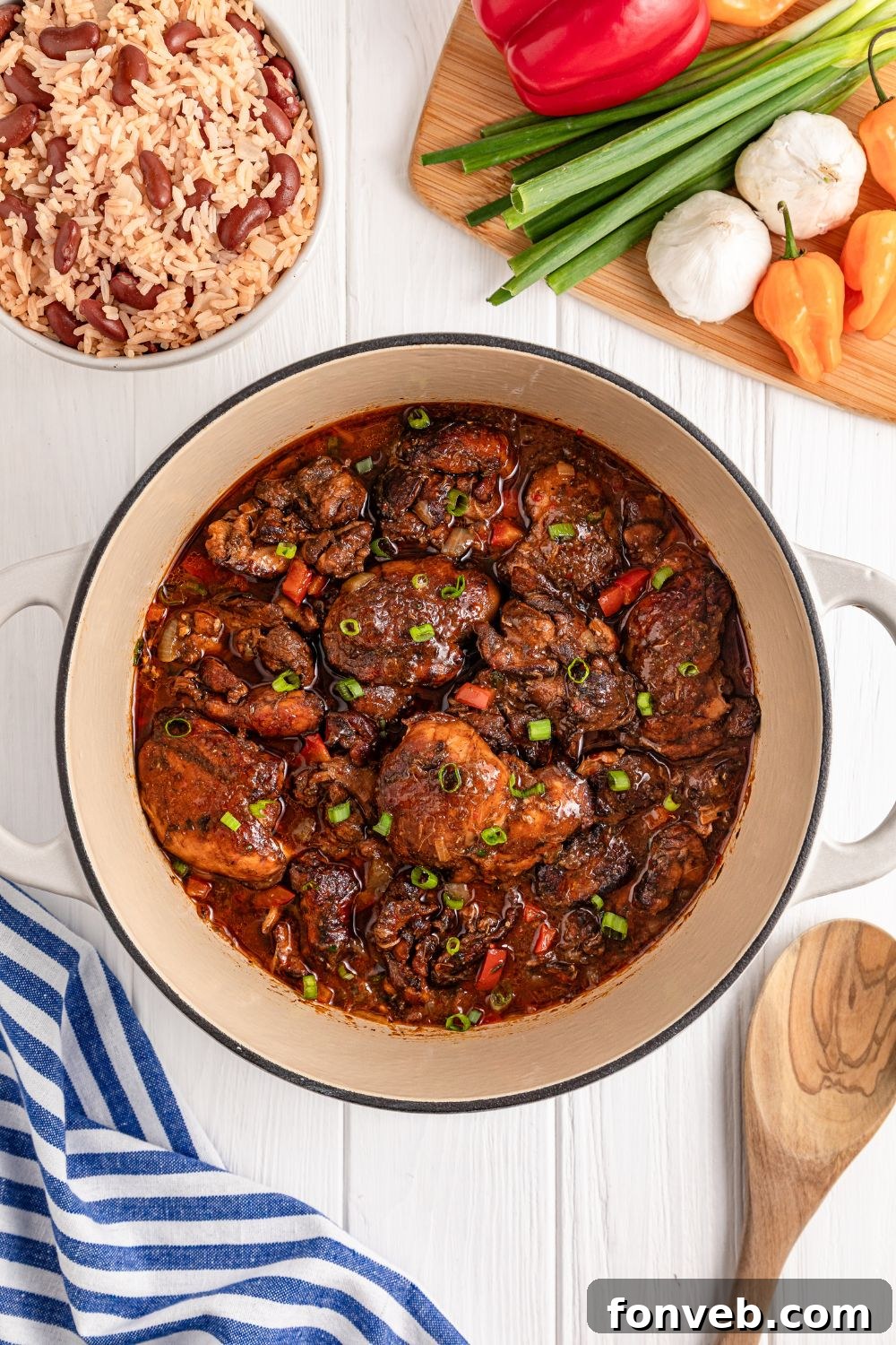 Jamaican Brown Stew Chicken in Dutch Oven sitting on table with rice and beans to side and a mix of vegetables 