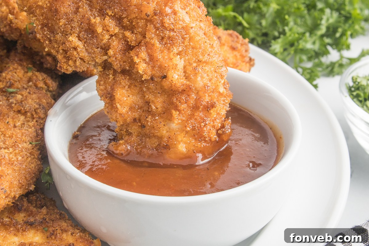 Ultra Crispy Air Fryer Chicken Tenders 13 A close-up shot of a crispy Air Fryer Chicken Tender being dipped into a small bowl of rich BBQ sauce