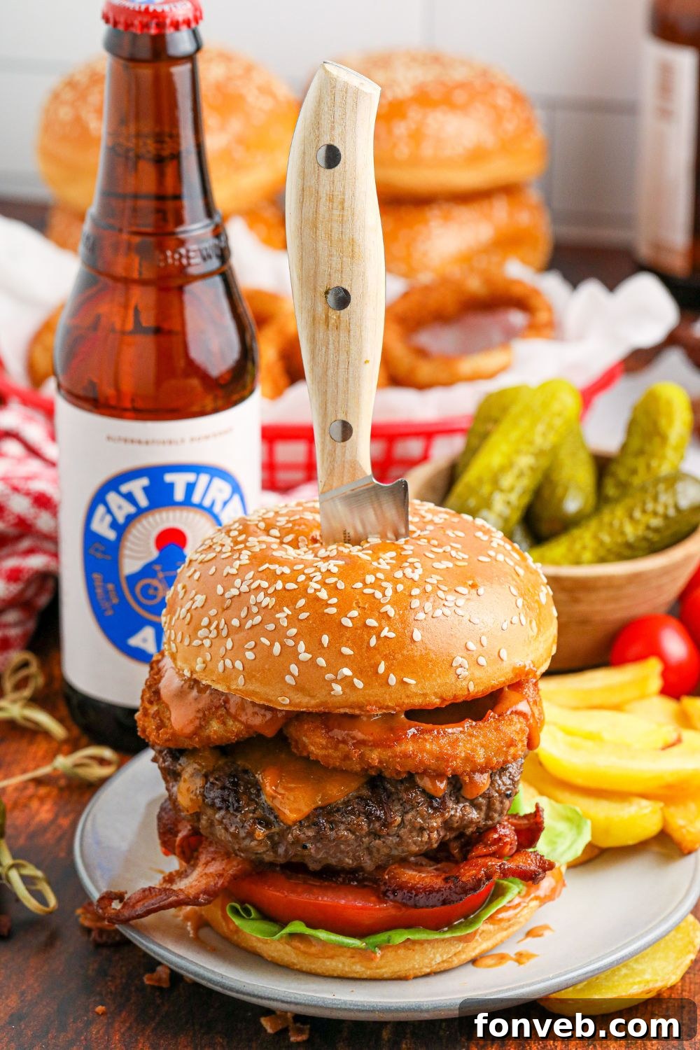 Chuck Wagon Burger 15 A Cowboy Burger with a knife stabbed into the center, indicating its height and deliciousness, with beer bottles and pickles blurred in the background.