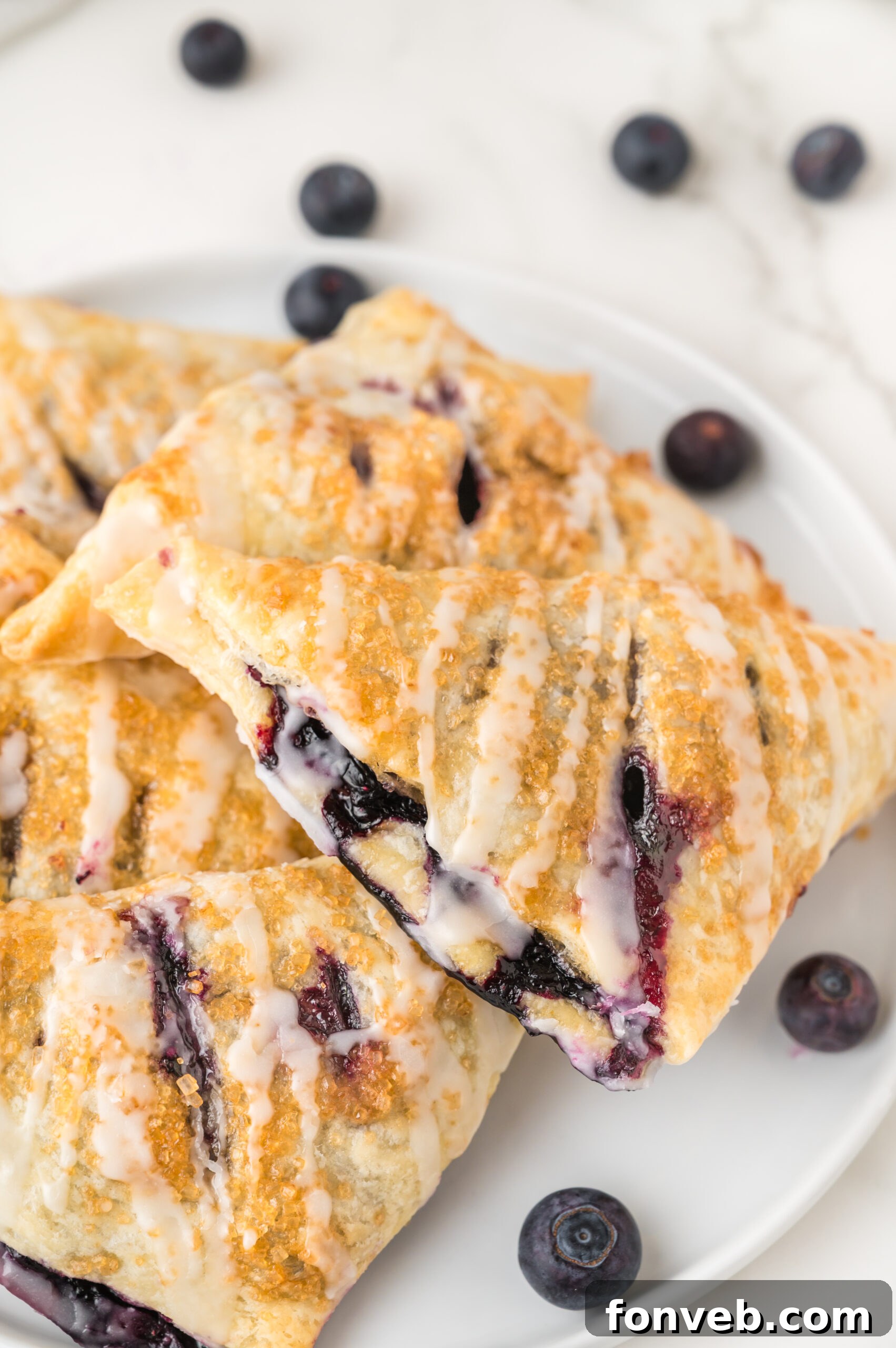 Freshly baked blueberry turnovers arranged artfully on a white plate, surrounded by ripe, glistening fresh blueberries, showcasing their golden crust and enticing filling.