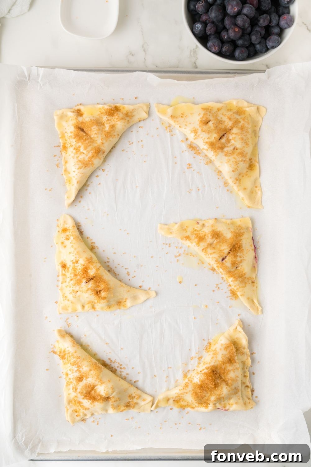 Blueberry turnovers on a baking tray, already brushed with butter and sprinkled with sugar, glistening and poised to go into the oven.