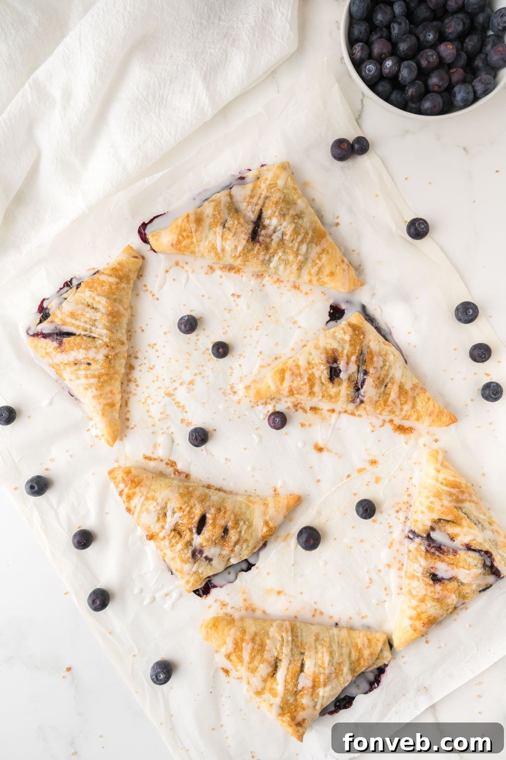Overhead view of perfectly baked blueberry turnovers on a baking tray, lightly drizzled with a sweet glaze, highlighting their golden crust and enticing finish.