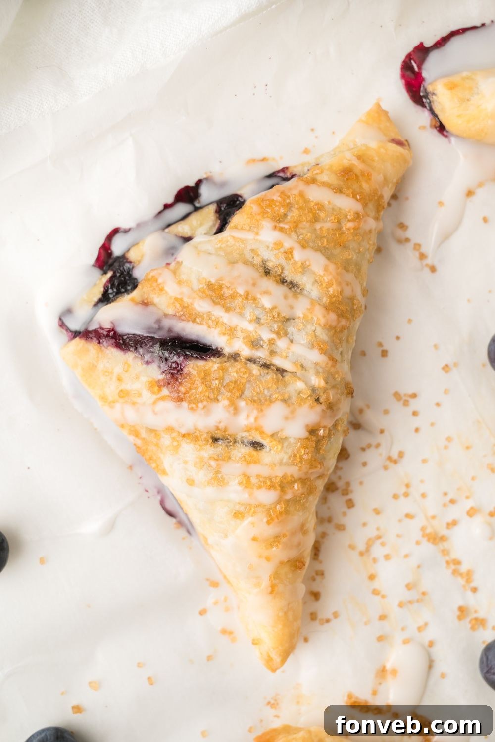 Overhead shot of a single golden blueberry turnover resting on parchment paper, displaying its flaky crust and delicate sugar crystals.