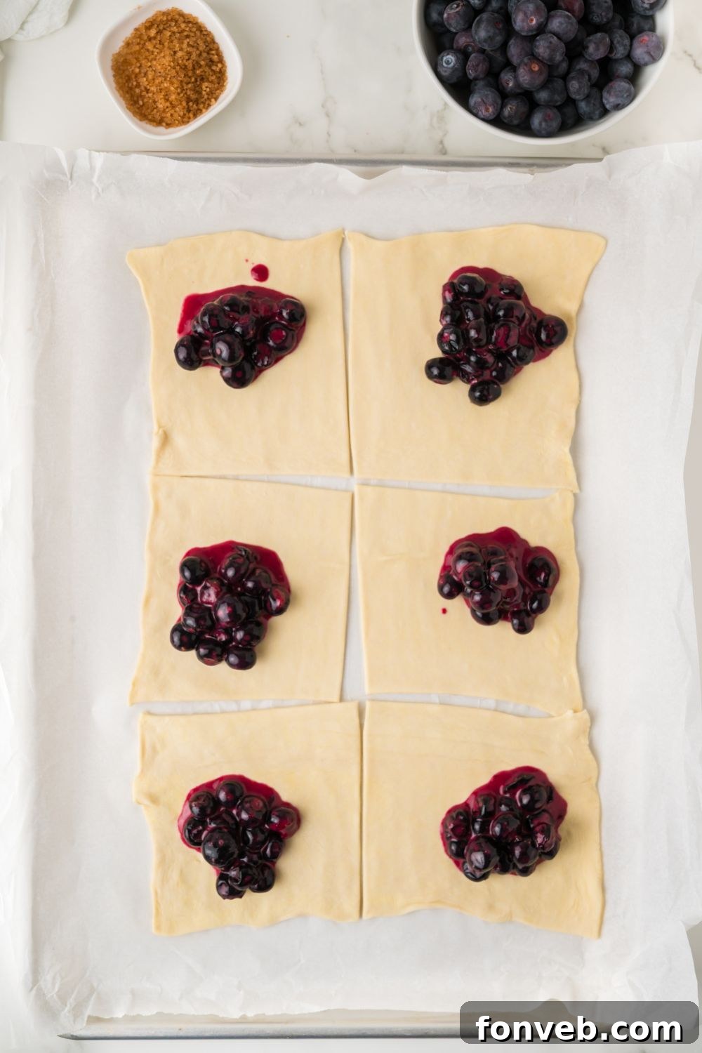 Puff pastry squares laid out, each generously topped with a spoonful of rich blueberry filling, prepared for folding into turnovers.