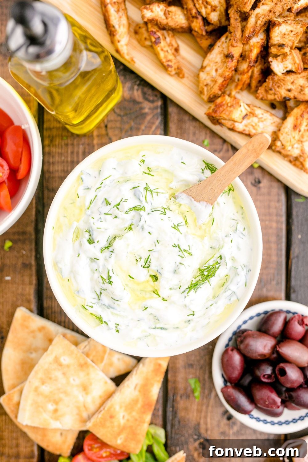 Tzatziki Sauce with a spoon in bowl and chicken and other ingredients around the bowl