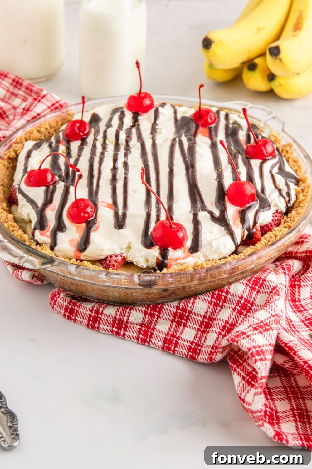 A No Bake Banana Split Pie displayed on a table, flanked by glasses of milk and fresh bananas arranged behind it, creating a rustic yet inviting scene.