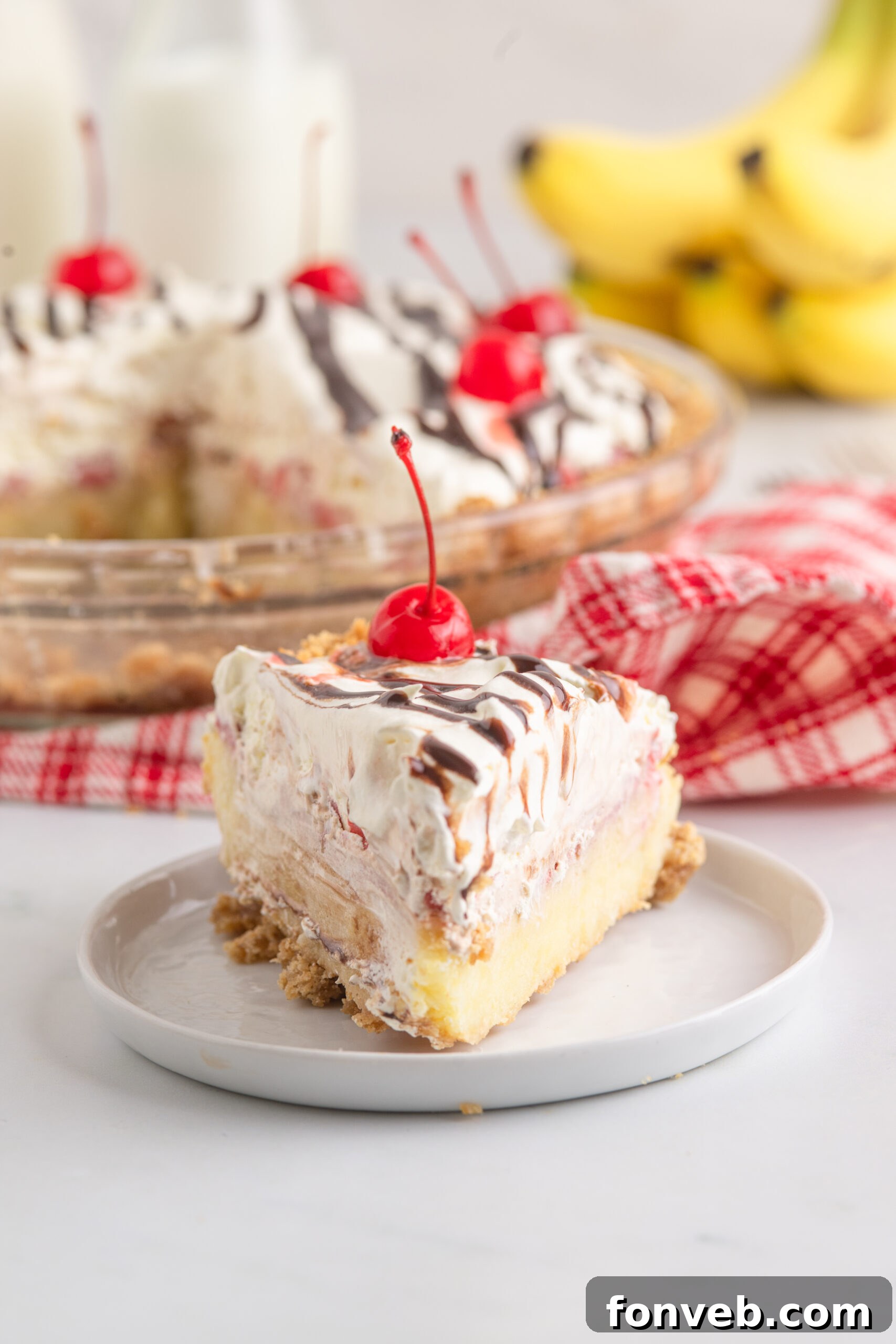 A delectable slice of No Bake Banana Split Pie on a white plate, with the remainder of the whole pie artfully blurred in the background.