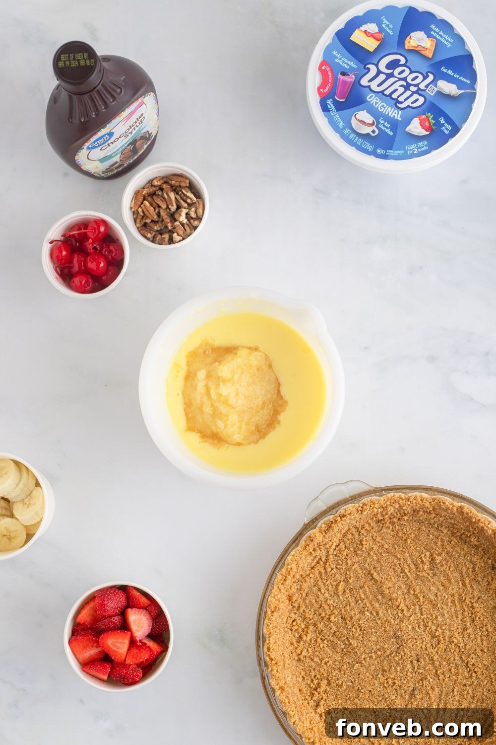 An array of ingredients for the No Bake Banana Split Pie including a bowl of pudding, a prepared crust, and various fresh fruits.