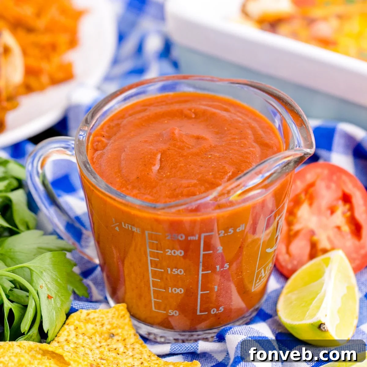Homemade Enchilada Sauce in a measuring cup on table with tomato, lemon wedge and more around container