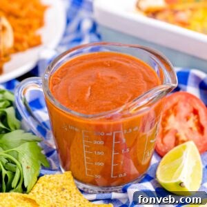 Homemade Enchilada Sauce in a measuring cup on table with tomato, lemon wedge and more around container