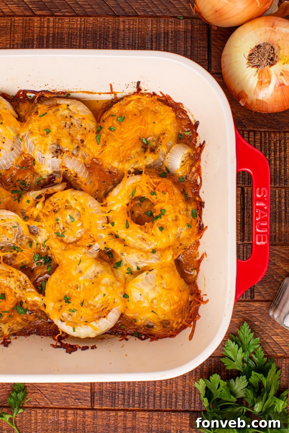 Staub pan on table with Cheesy onion bake in a pan with onions and parsley around the table