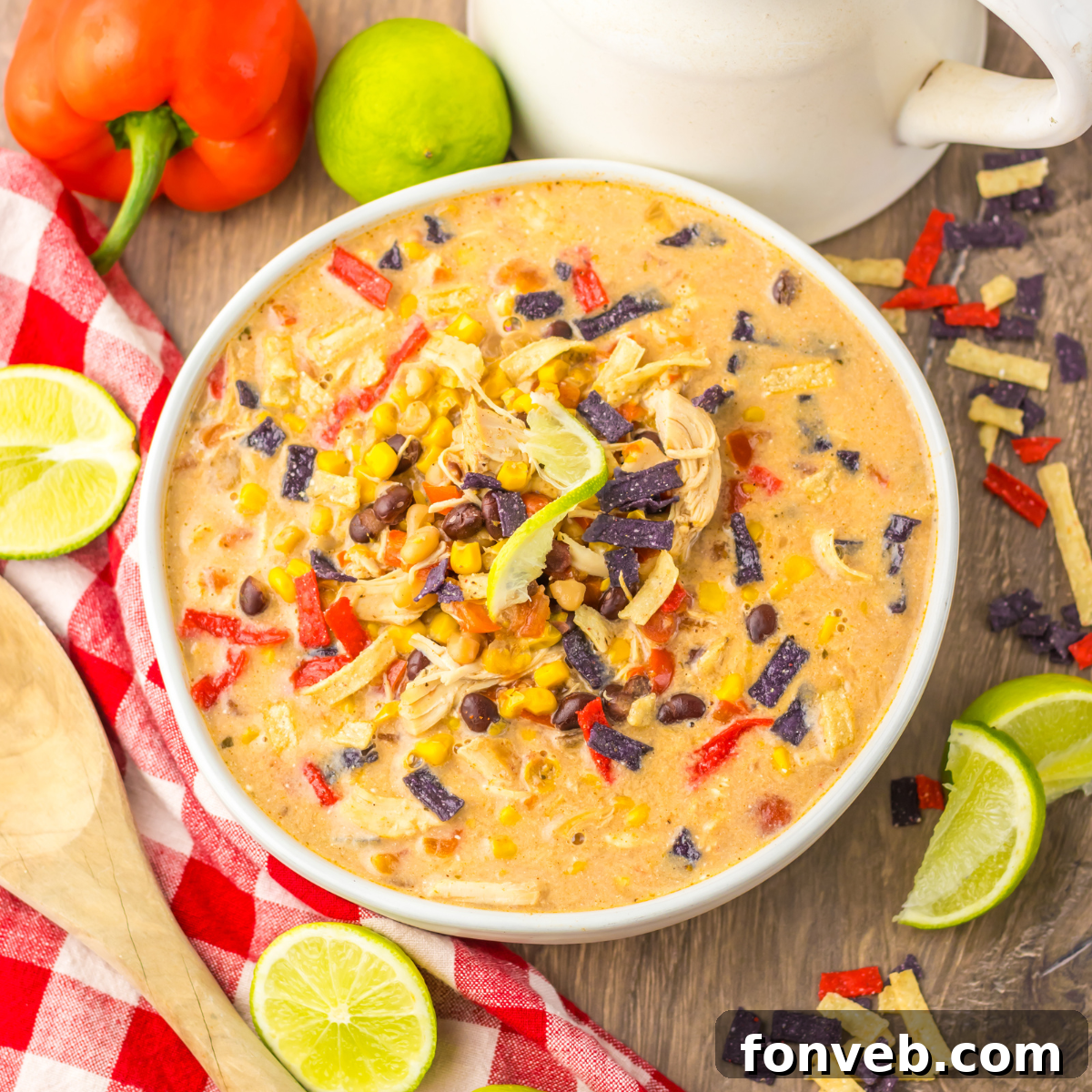 An overhead view of a rustic white bowl brimming with Slow Cooker Tortilla Soup, artfully placed beside a white and red checkered napkin and several slices of fresh lime.