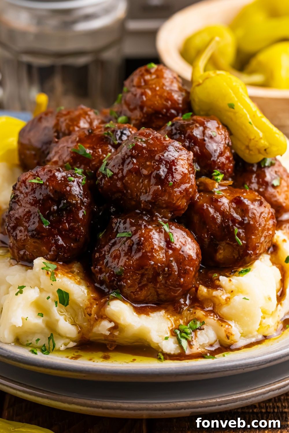 Crockpot Mississippi Meatballs piled in a stack on a plate with a bowl of peppers behind it on table and mashed potatoes on the plate with meatballs 
